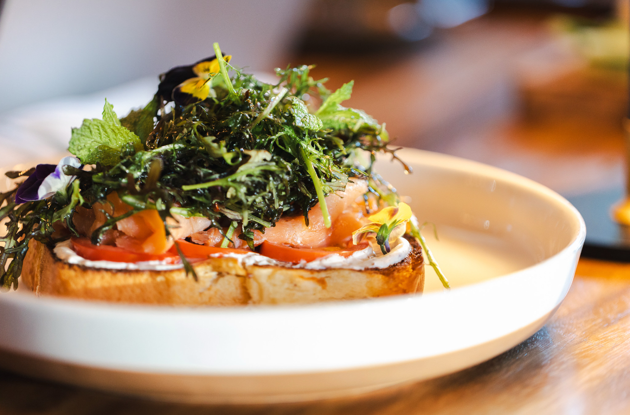 Smoked salmon tartine with tomato, crème fraîche, herb salad and edible flowers plated in a wide ceramic bowl